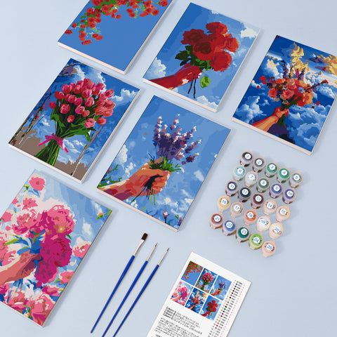 Six mini paint by numbers canvases arranged in a set, each featuring a vibrant floral bouquet illustration held in hand against a bright blue sky background. The set includes red roses, pink tulips, purple lavender, and mixed wildflower arrangements, all painted in a bold block-style illustration with strong color contrast, simplified brush textures, and expressive painterly details.